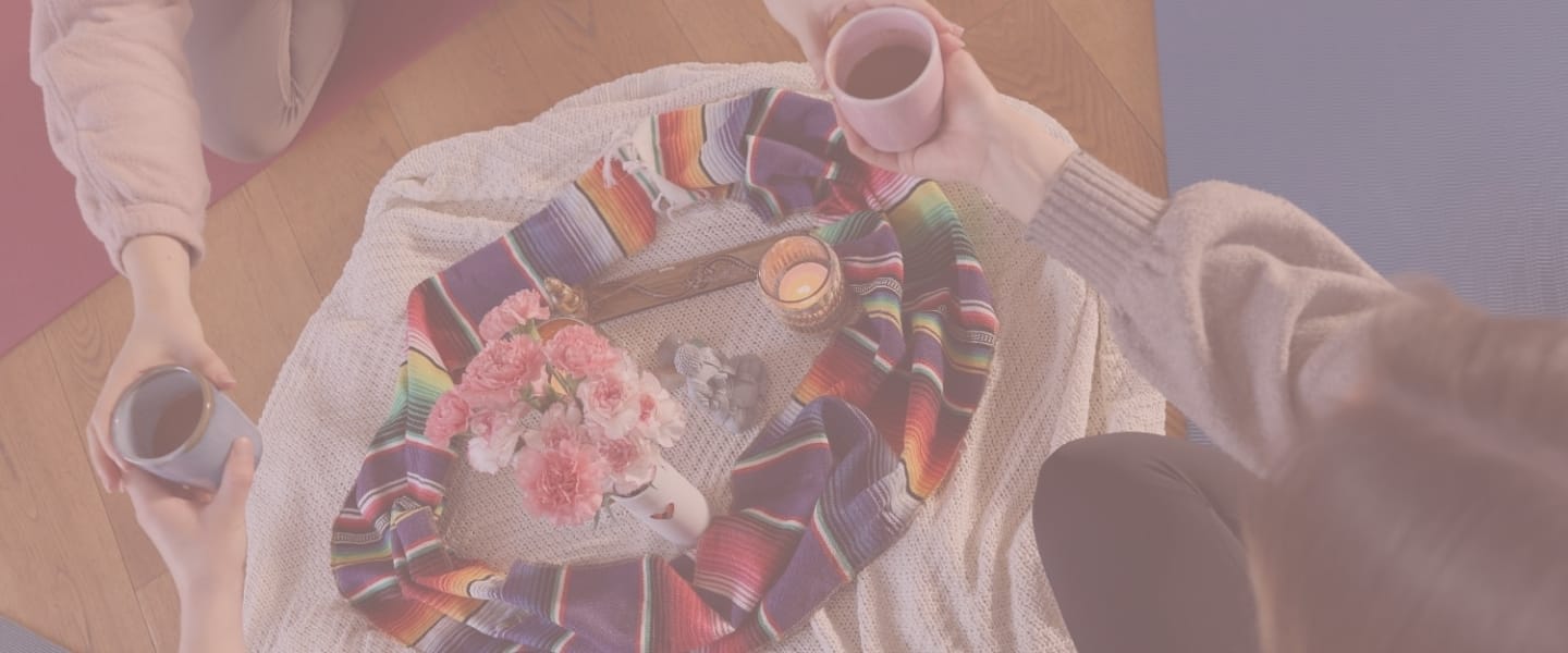 Ansicht von oben auf eine kleine Gruppe von Personen, die auf dem Boden sitzen. Zwei Personen halten Becher mit einem dunklen Getränk in den Händen und reichen sie einander. In der Mitte liegt eine weiße Decke, kreisförmig dekoriert mit einem bunten mexikanischen Tuch, einer Vase mit rosa Nelken, einer kleinen Buddha-Statue und einer brennenden Kerze im Glas. Das Bild hat einen zartrosa Farbfilter.