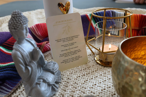 Nahaufnahme einer Impulskarte mit der Überschrift „Leichtigkeit“ und Reflexionsfragen, platziert neben einer Buddha-Figur, einem brennenden Teelicht im goldenen Halter und einer Opferschale auf einer hellen Strickdecke.
