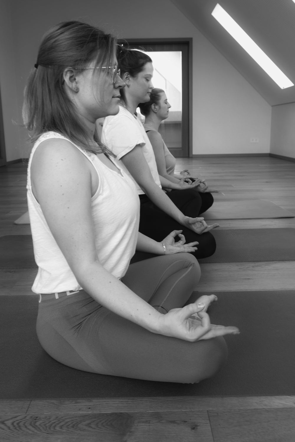 Schwarz-weiß-Aufnahme von drei Frauen, die hintereinander im Meditationssitz auf Yoga-Matten sitzen, die Augen geschlossen haben und ihre Hände im Chin-Mudra halten.