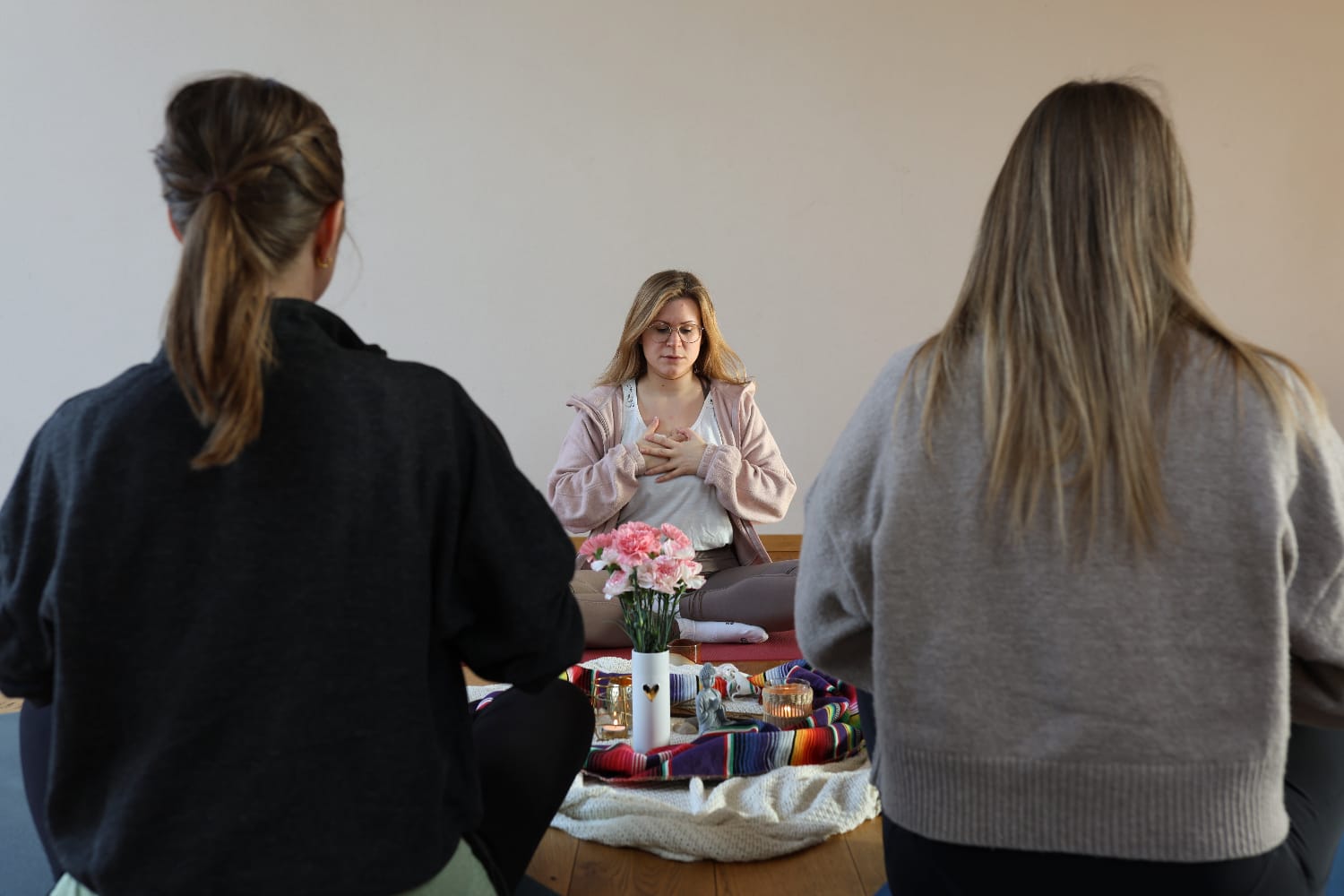 Eine Frau mit geschlossenen Augen sitzt meditierend im Zentrum einer Gruppe, die Hände auf das Herz gelegt, während zwei weitere Personen ihr im Vordergrund gegenübersitzen.