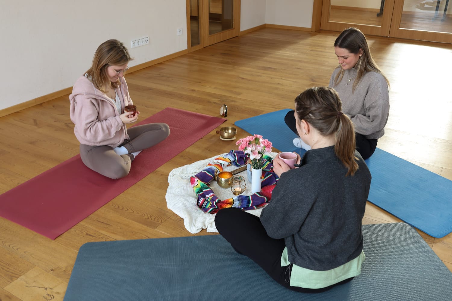 Drei Frauen sitzen im Kreis auf bunten Yoga-Matten um einen rituellen Altar mit Blumen, Kerzen und einer Klangschale, während sie achtsam aus ihren Tassen trinken.