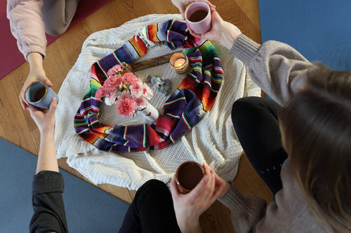 Vogelperspektive auf drei Frauen, die im Kreis um einen geschmückten Altar mit Blumen und Kerzen sitzen und gemeinsam aus Keramiktassen trinken.