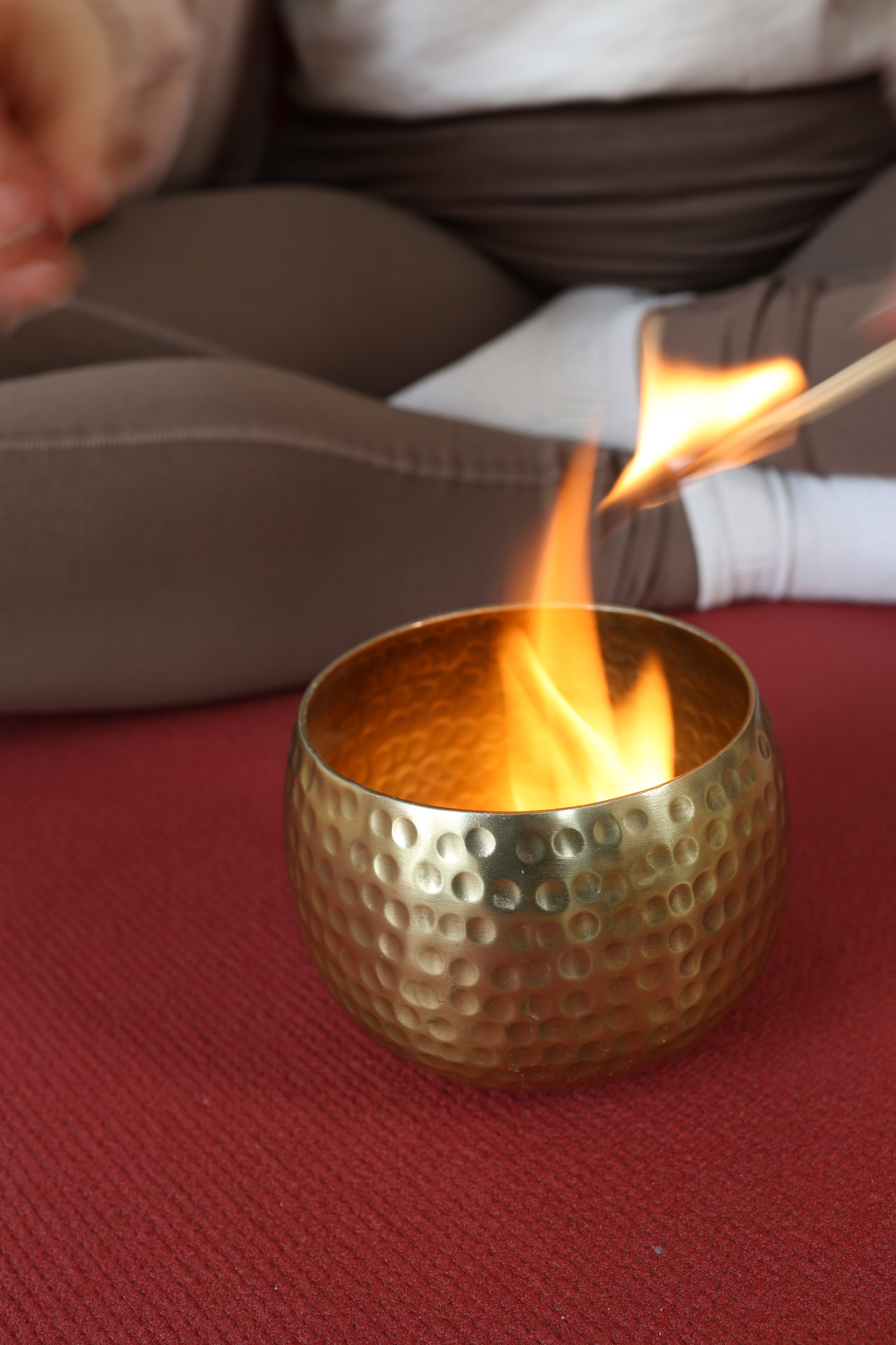 Nahaufnahme einer gehämmerten Messingschale mit einer brennenden Flamme auf einer roten Yoga-Matte, im Hintergrund eine Person im Meditationssitz.