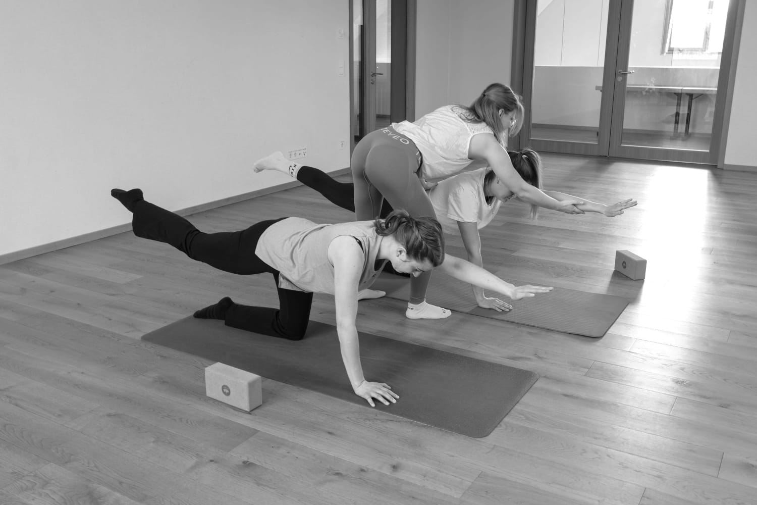 Schwarz-weiß-Foto von drei Frauen, die auf Yoga-Matten trainieren, während eine Trainerin eine Teilnehmerin in der Bird-Dog-Pose korrigiert.